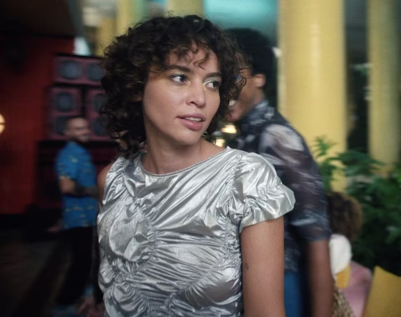 Una mujer con el pelo rizado y una blusa plateada brillante se encuentra en un animado interior con columnas amarillas y plantas. En el fondo, la gente socializa, creando un ambiente vibrante.