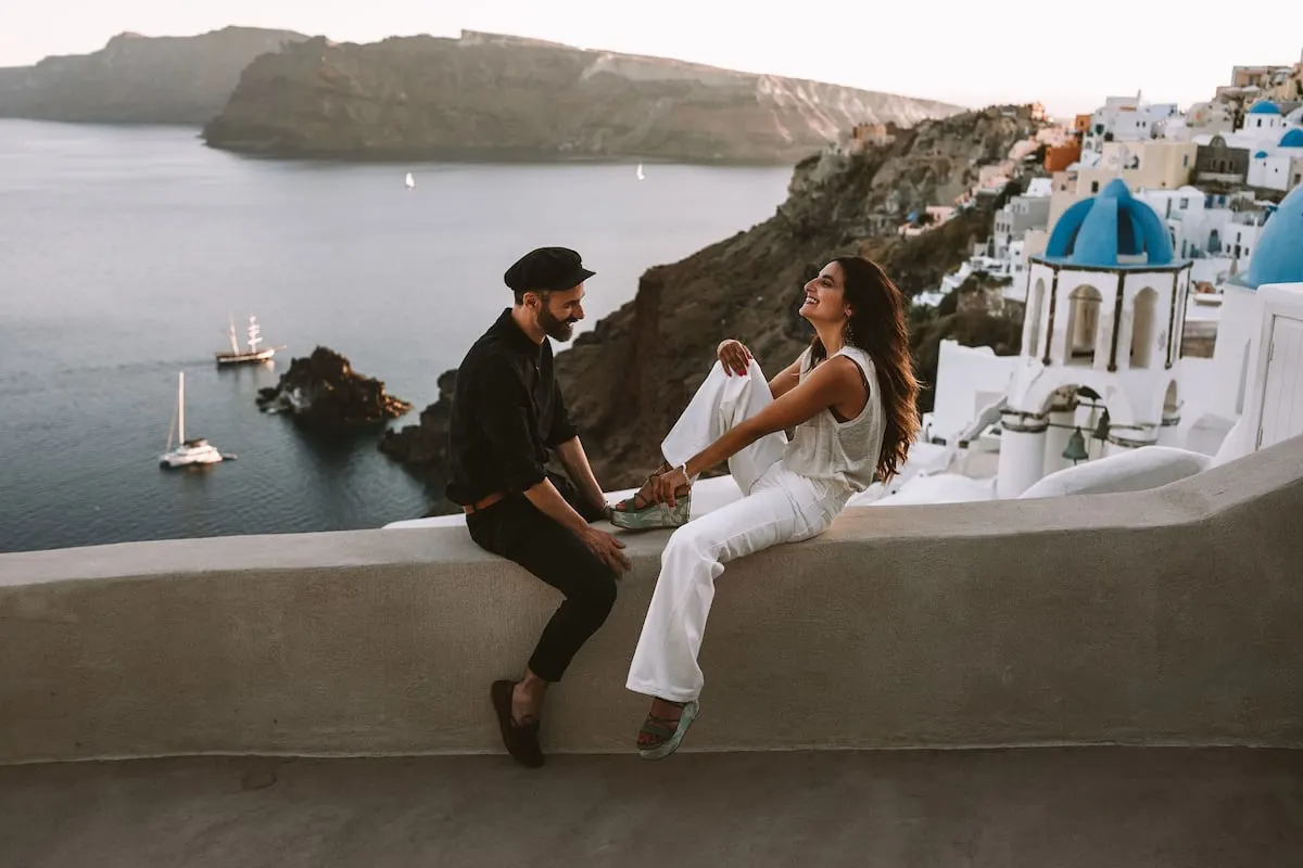 couple perched on wall in greece above city and ocean