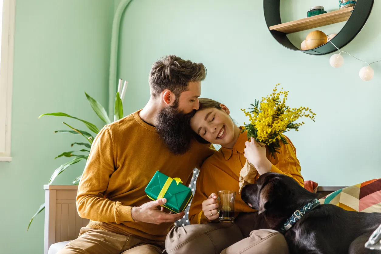 man kisses woman who is holding a flower bouquet while their dog cuddles them