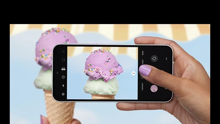 Una mano tomando una foto con un dispositivo Galaxy S23 FE visto desde el frente y horizontalmente. La pantalla en modo de imagen muestra una imagen clara y detallada de un cucharón de helado en primer plano con pepitas de colores.