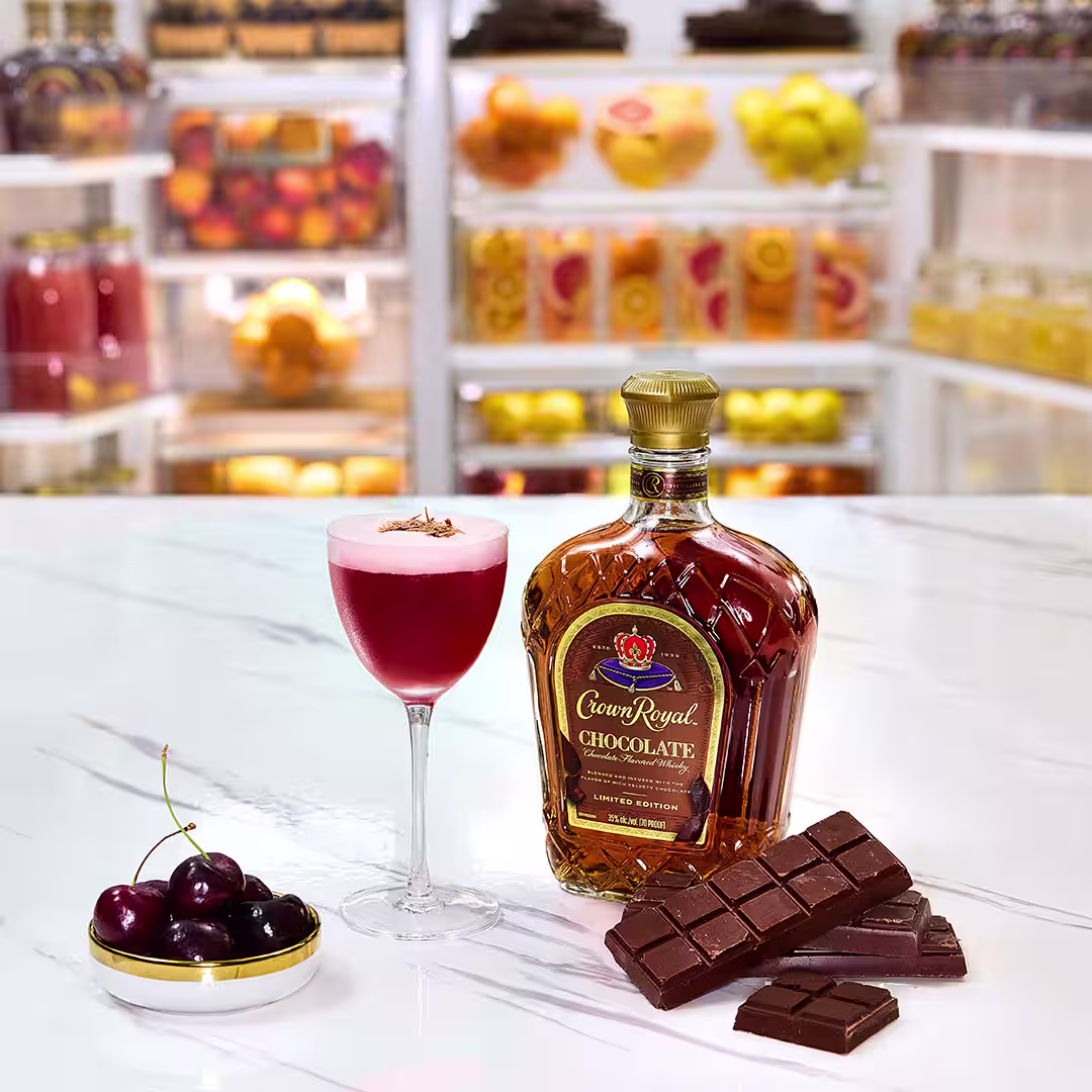 A bottle of Crown Royal Chocolate next to a chocolate cherry sour cocktail, a plate of cherries and a chocolate bar