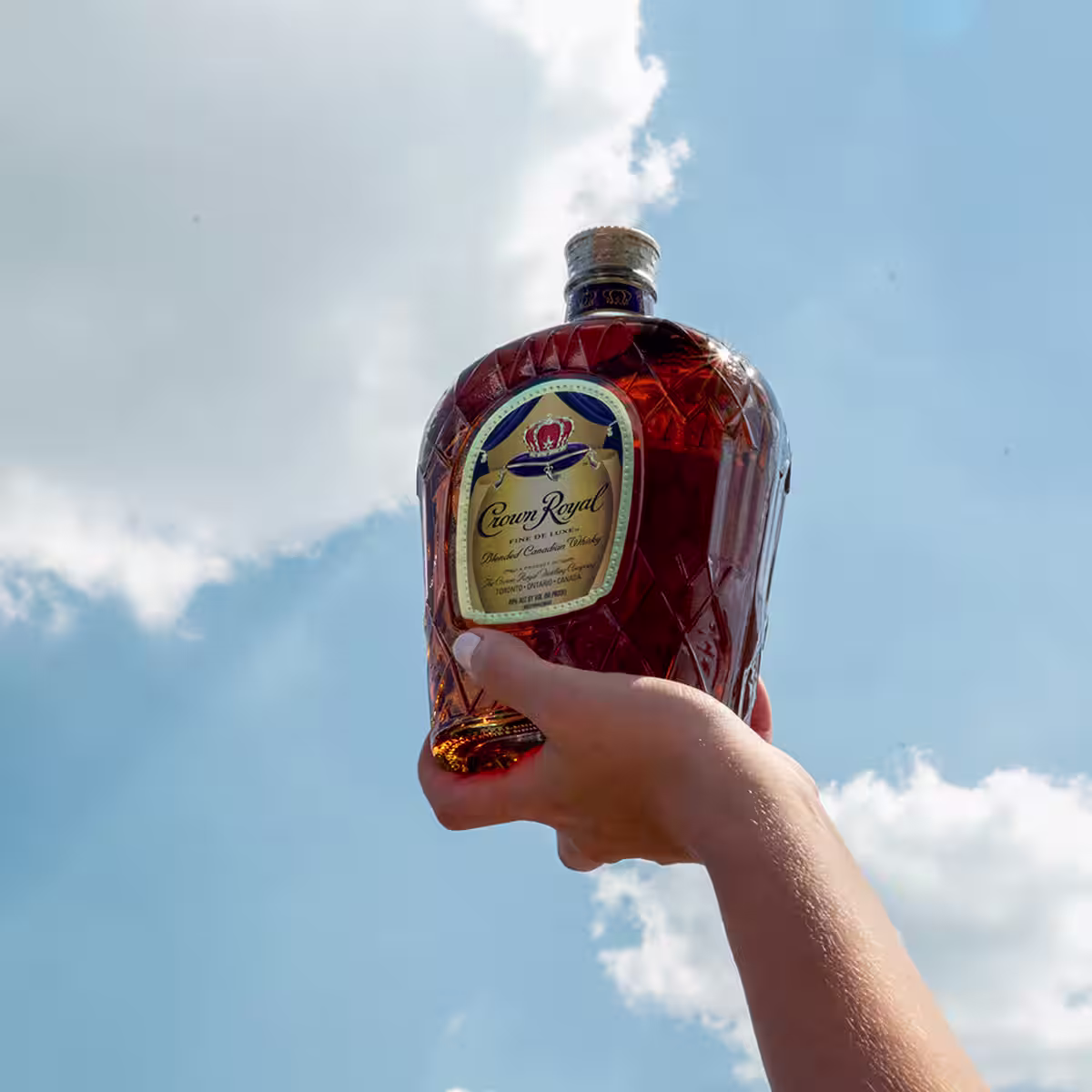 A hand holding a bottle of Crown Royal De Luxe against a blue sky