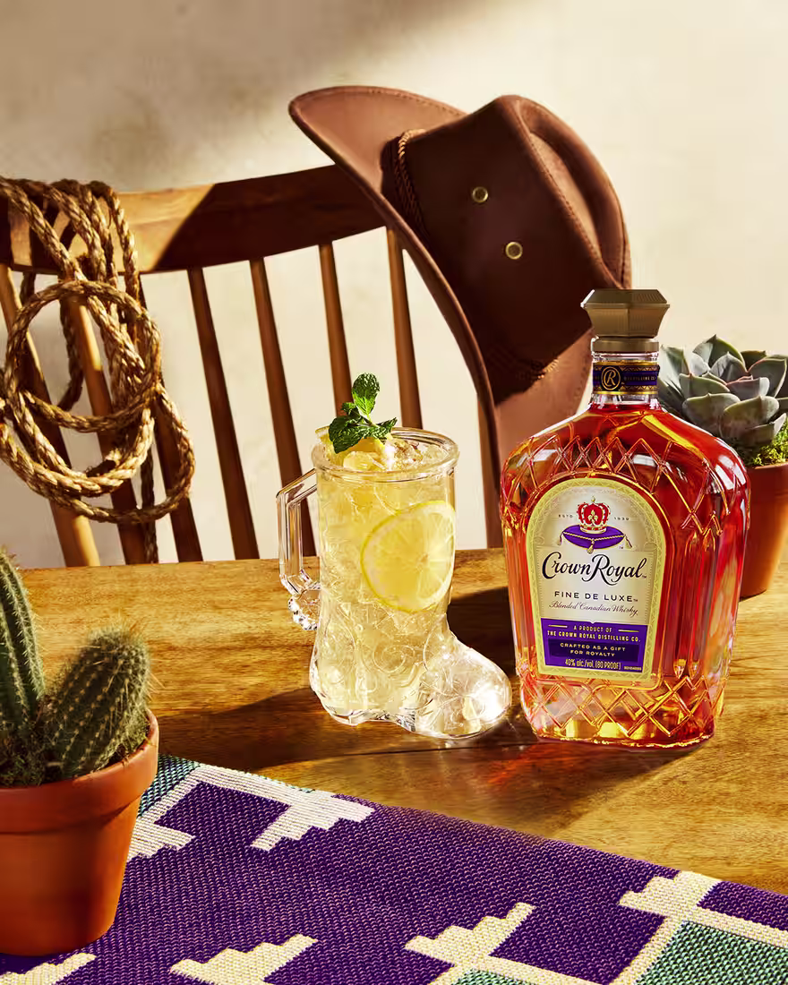 A yellow Royal Rider cocktail sitting on a table next to a bottle of Crown Royal Fine De Luxe. Around the cocktail is a plate of chips and salsa.