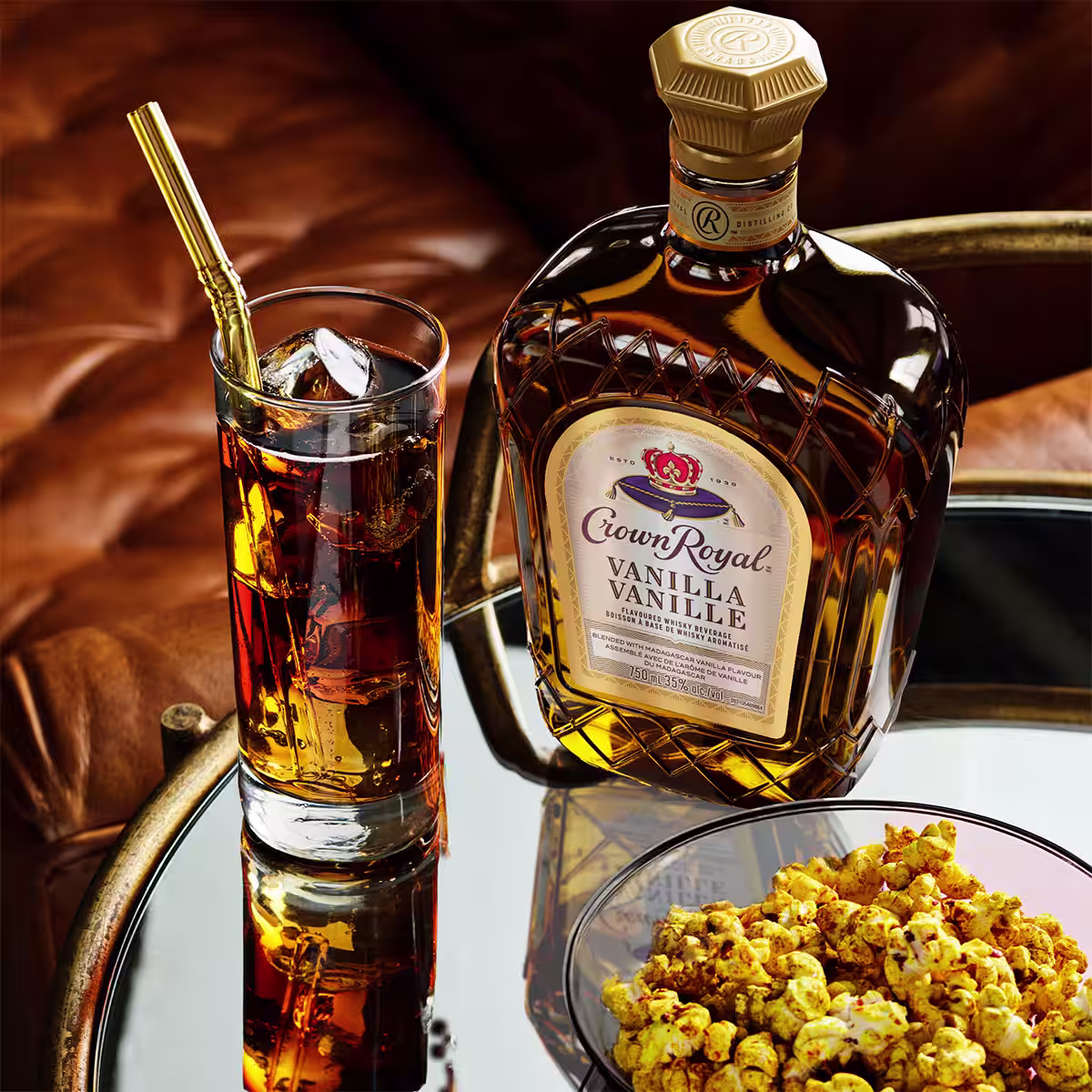 A bottle of Crown Royal Vanilla sitting next to a Vanilla & Cola cocktail in front of a bowl of popcorn