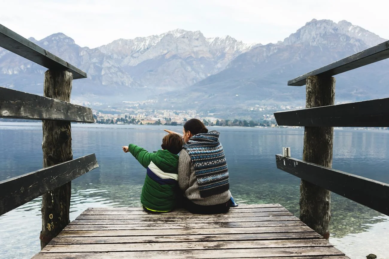 Child and a parent sitting on the pier, watching towards lake and mountains.