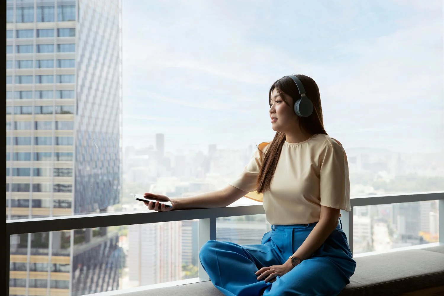 A woman sitting by a window holding a mobile phone listening to music