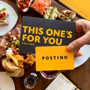 A hand holding cards that say "This One's For You Postino" in front of trays of food