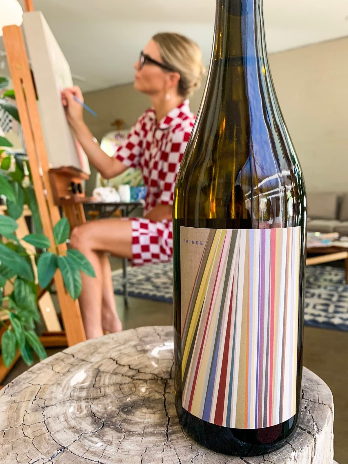 A woman painting at an easel with a wine bottle in the foreground