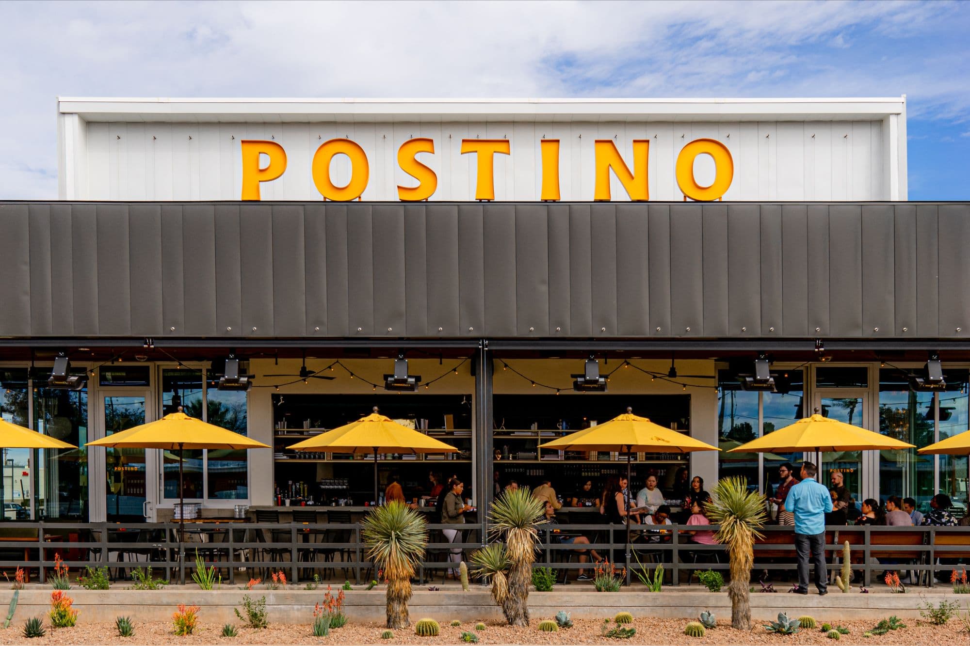 A restaurant with a large yellow Postino sign and umbrellas.
