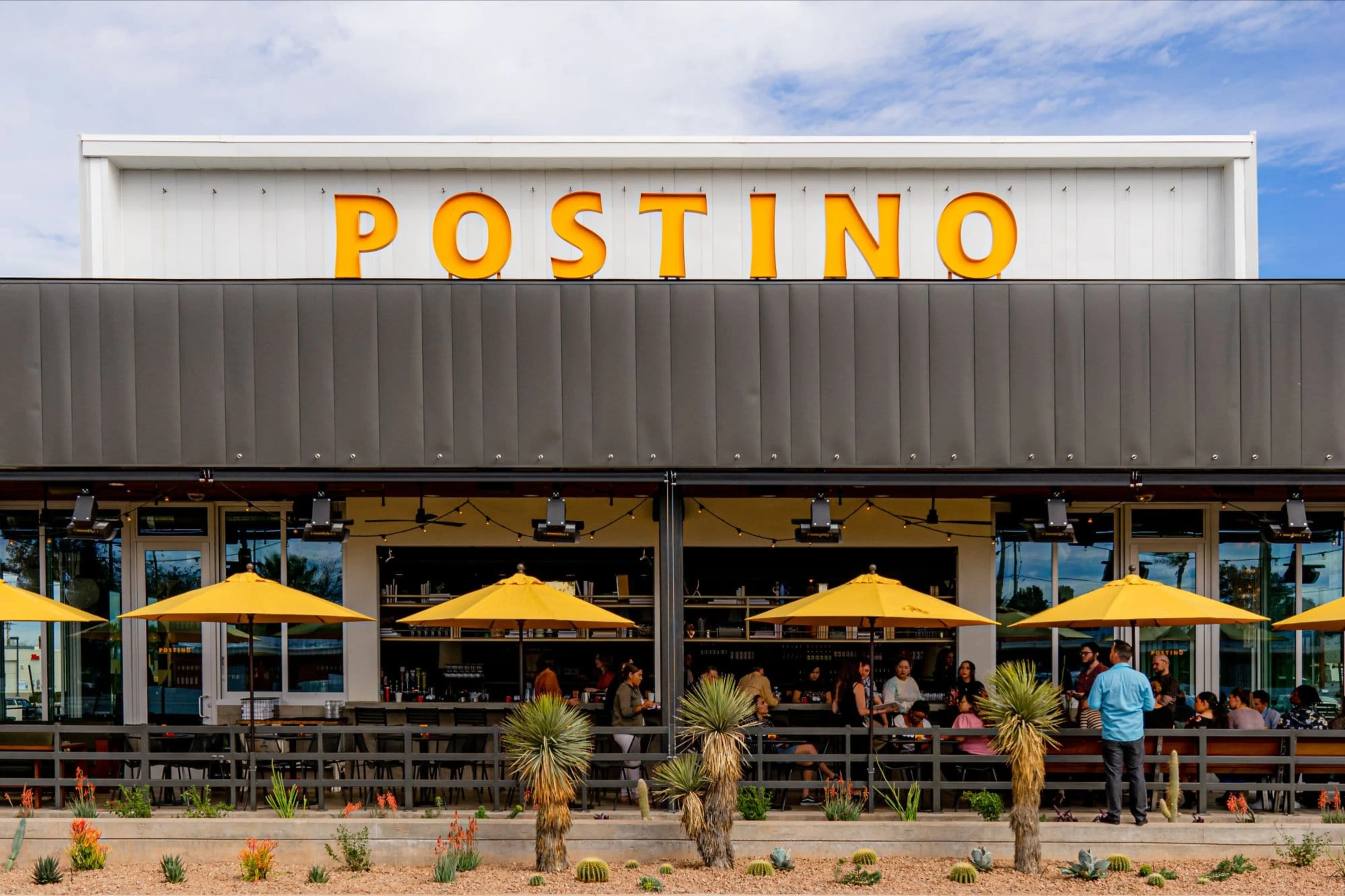 A restaurant with a large yellow Postino sign and umbrellas.