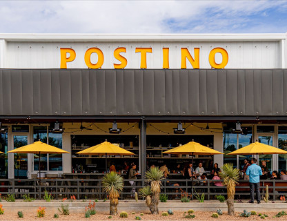 A restaurant with a large yellow Postino sign and umbrellas.