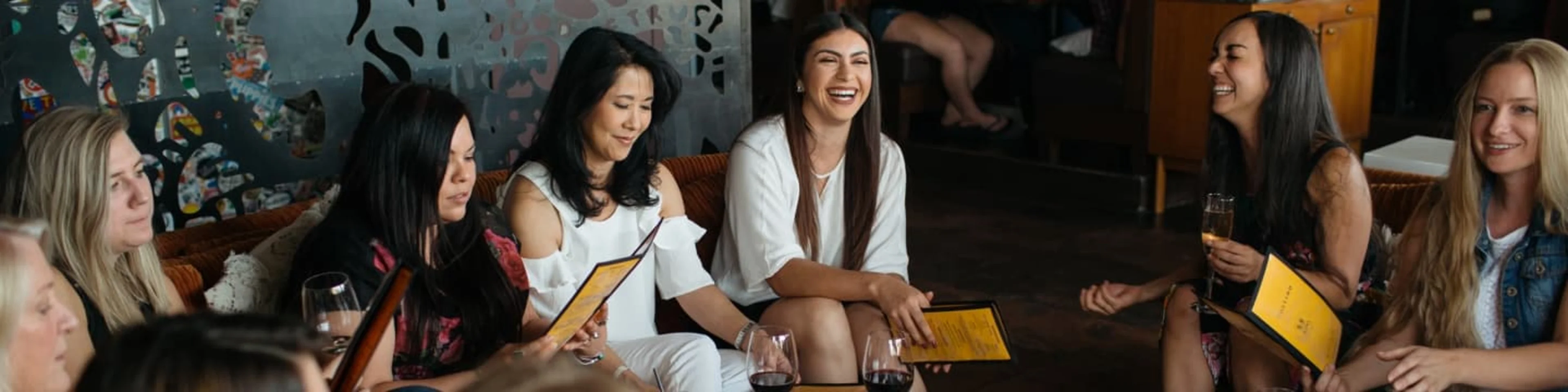 A group of women are seated at a table, laughing and holding menus.