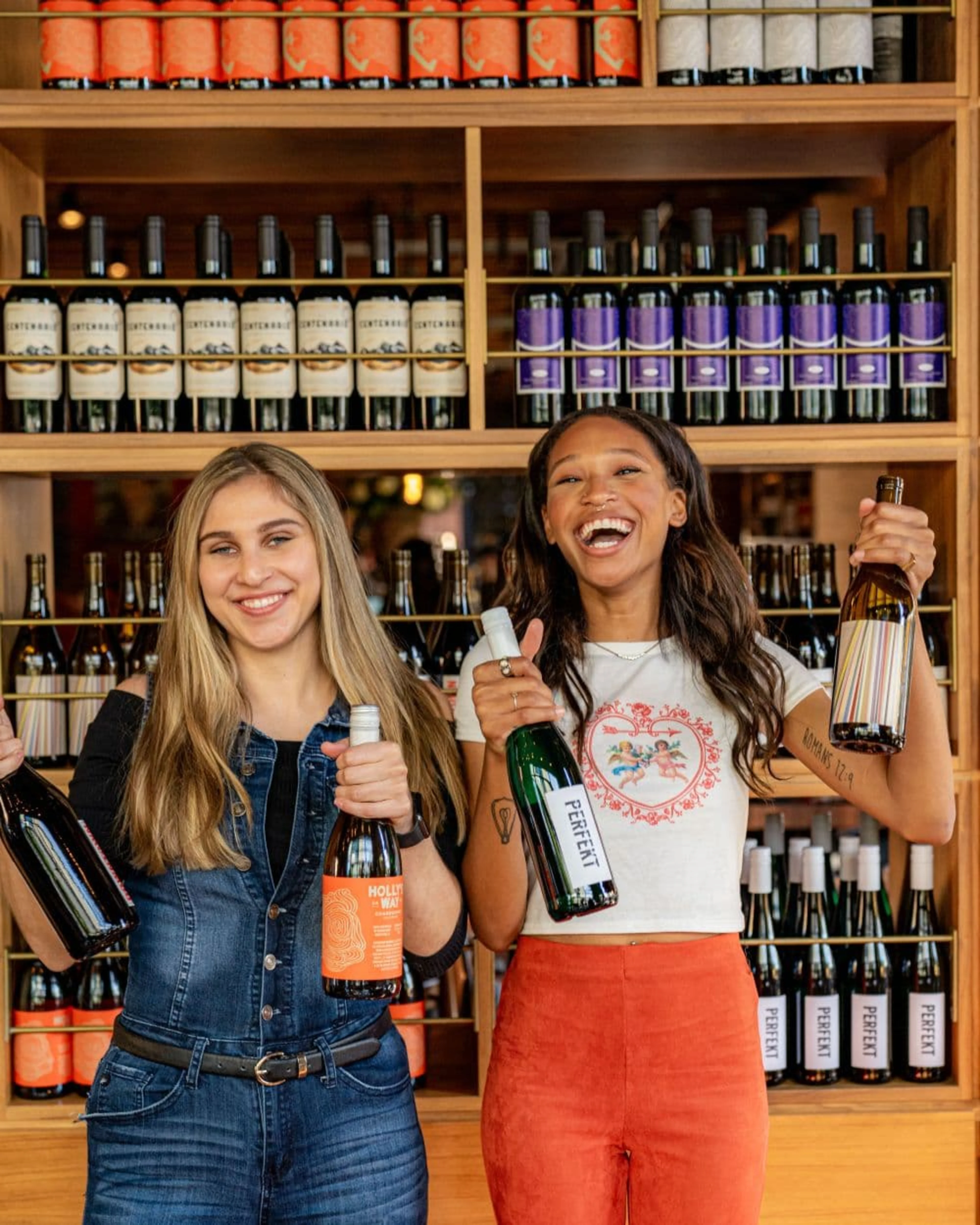 Two women smiling and holding up wine bottles in front of a wall of wine.
