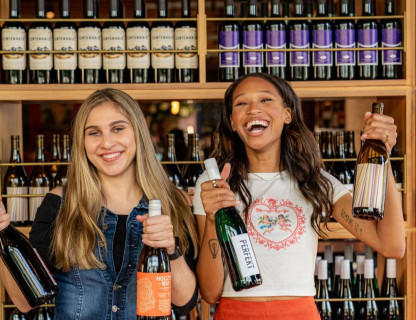 Two women smiling and holding up wine bottles in front of a wall of wine.