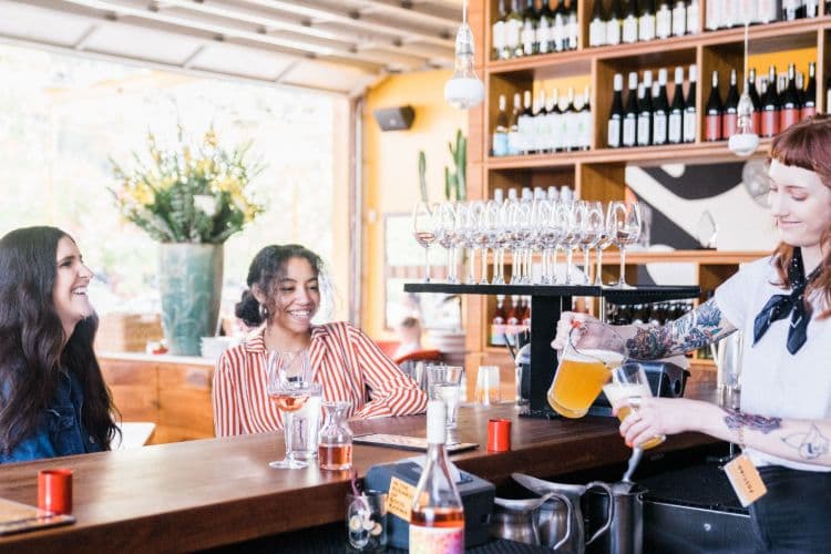 A bartender is pouring a pitcher of beer to two customers.