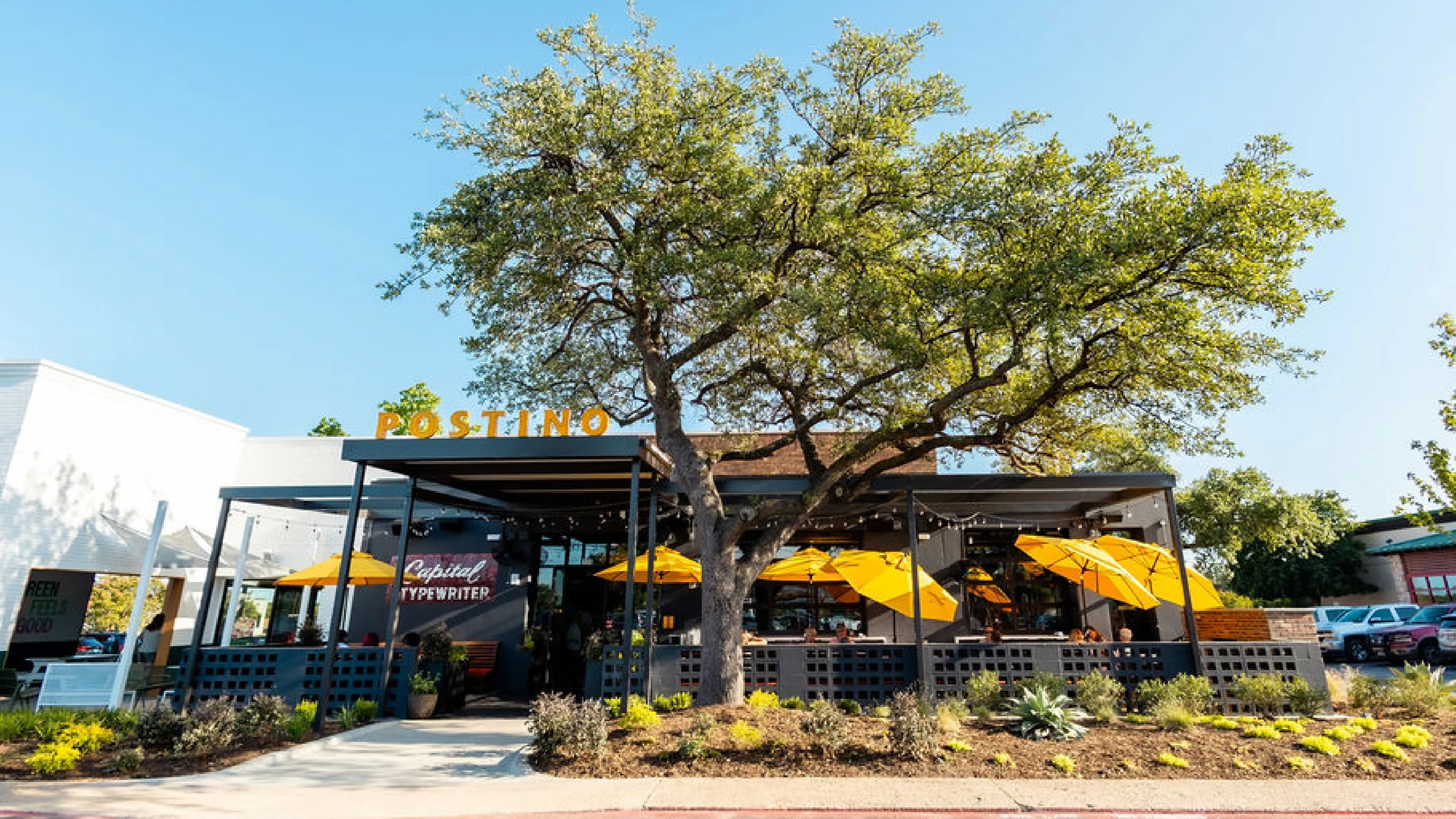 The exterior of Postino Addison with a bright yellow sign and a large tree. 