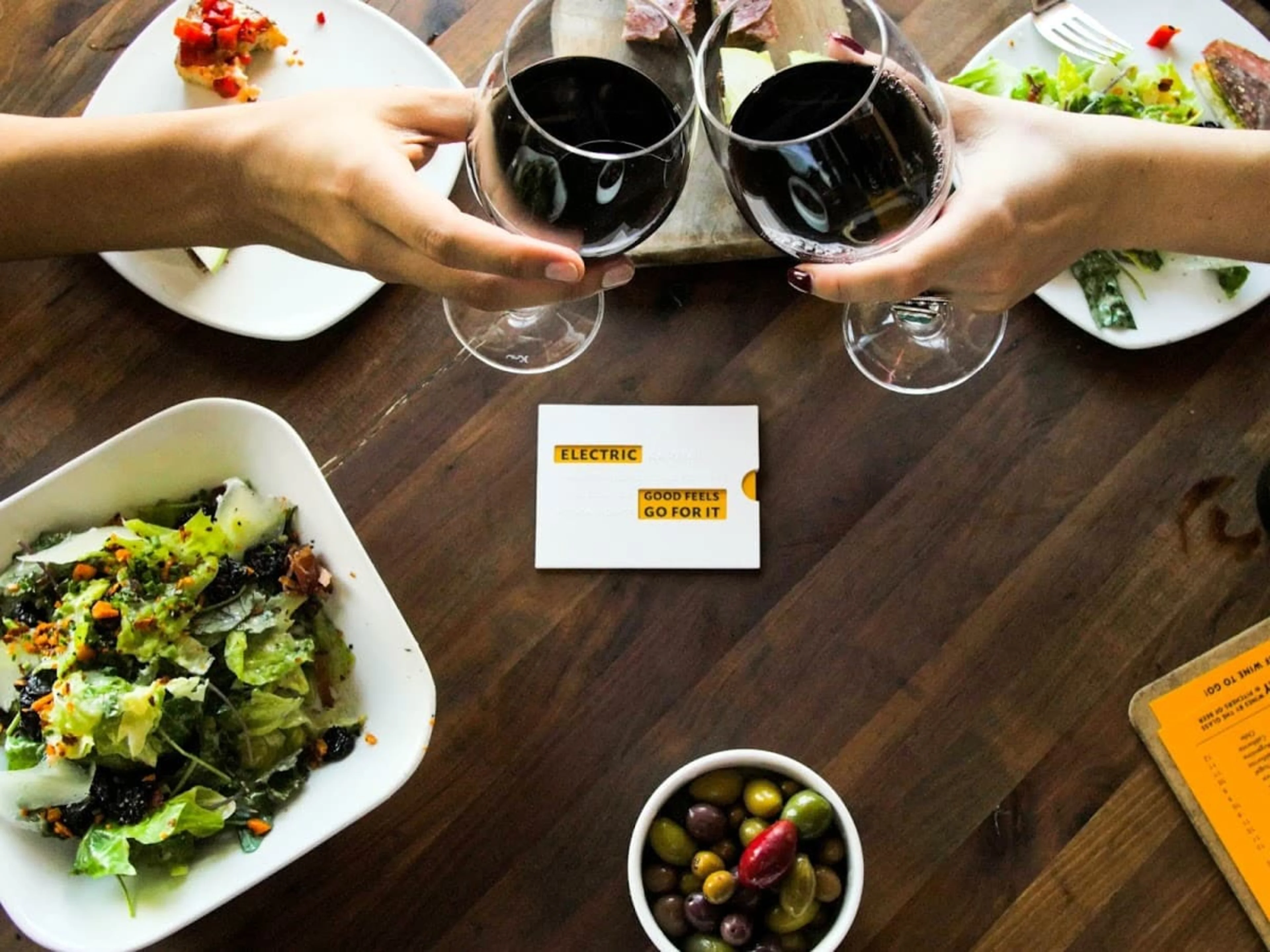 A wooden table with food platters and people clicking red wine glasses.