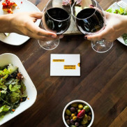 A wooden table with food platters and people clicking red wine glasses. 