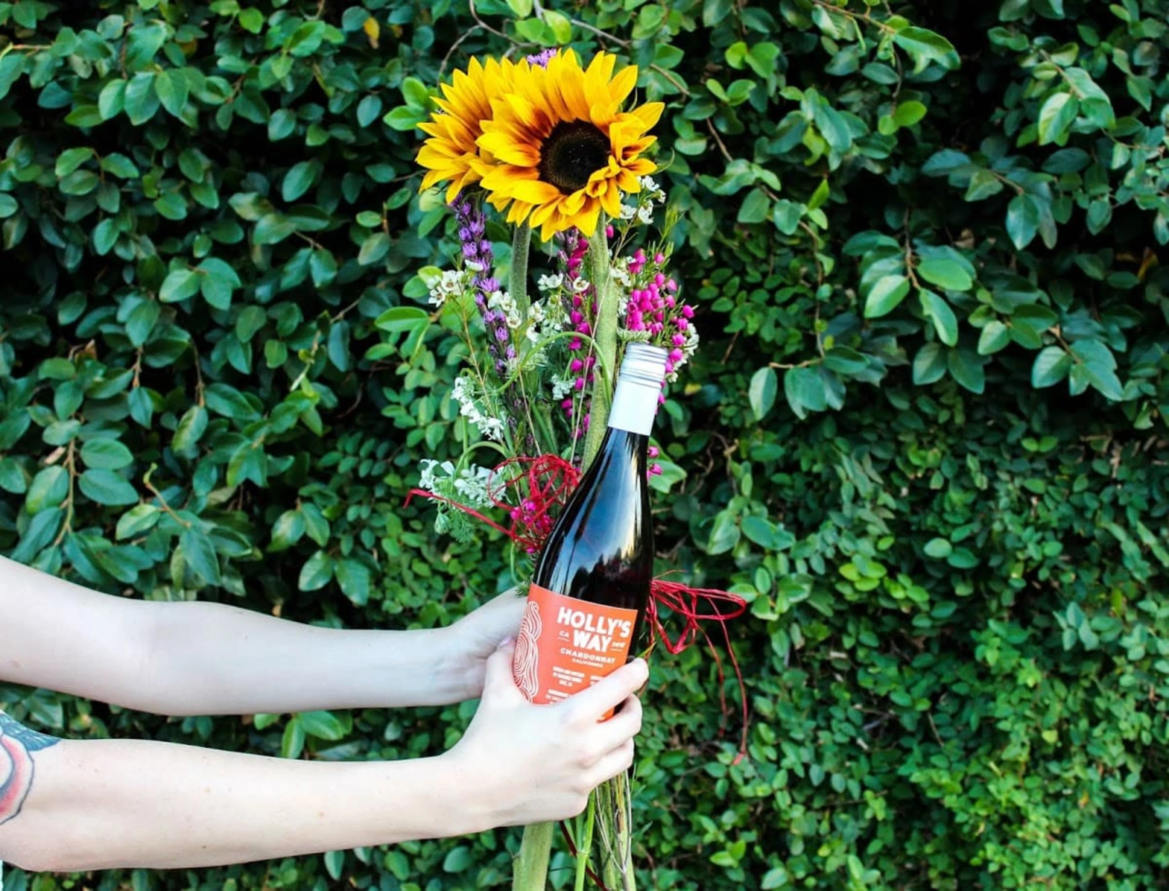 A person holding a bouquet of flowers and a bottle of wine in front of a shrub.