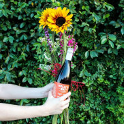 A person holding a bouquet of flowers and a bottle of wine in front of a shrub.
