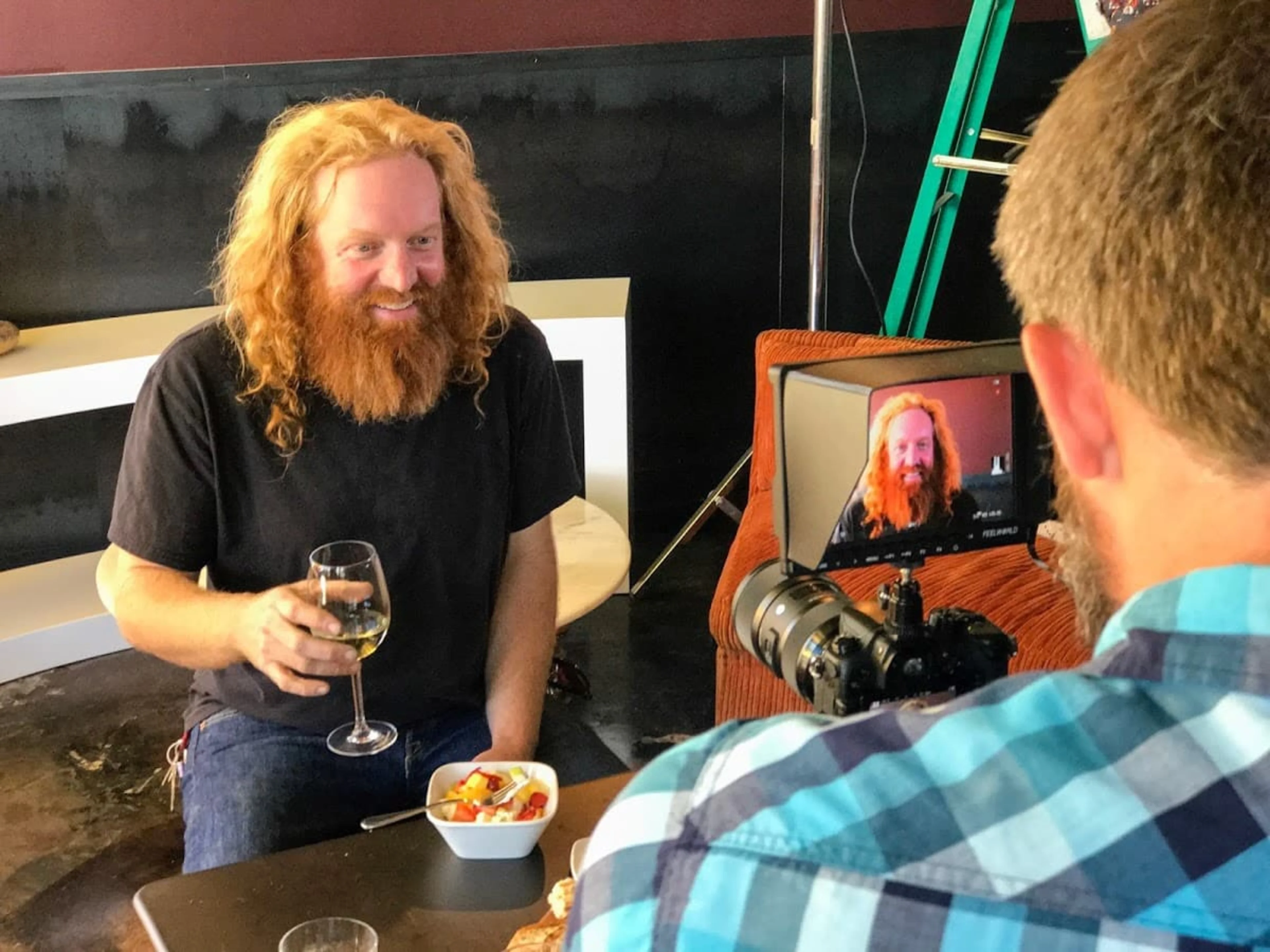 A red-headed man holding a glass of wine is being interviewed and filmed.