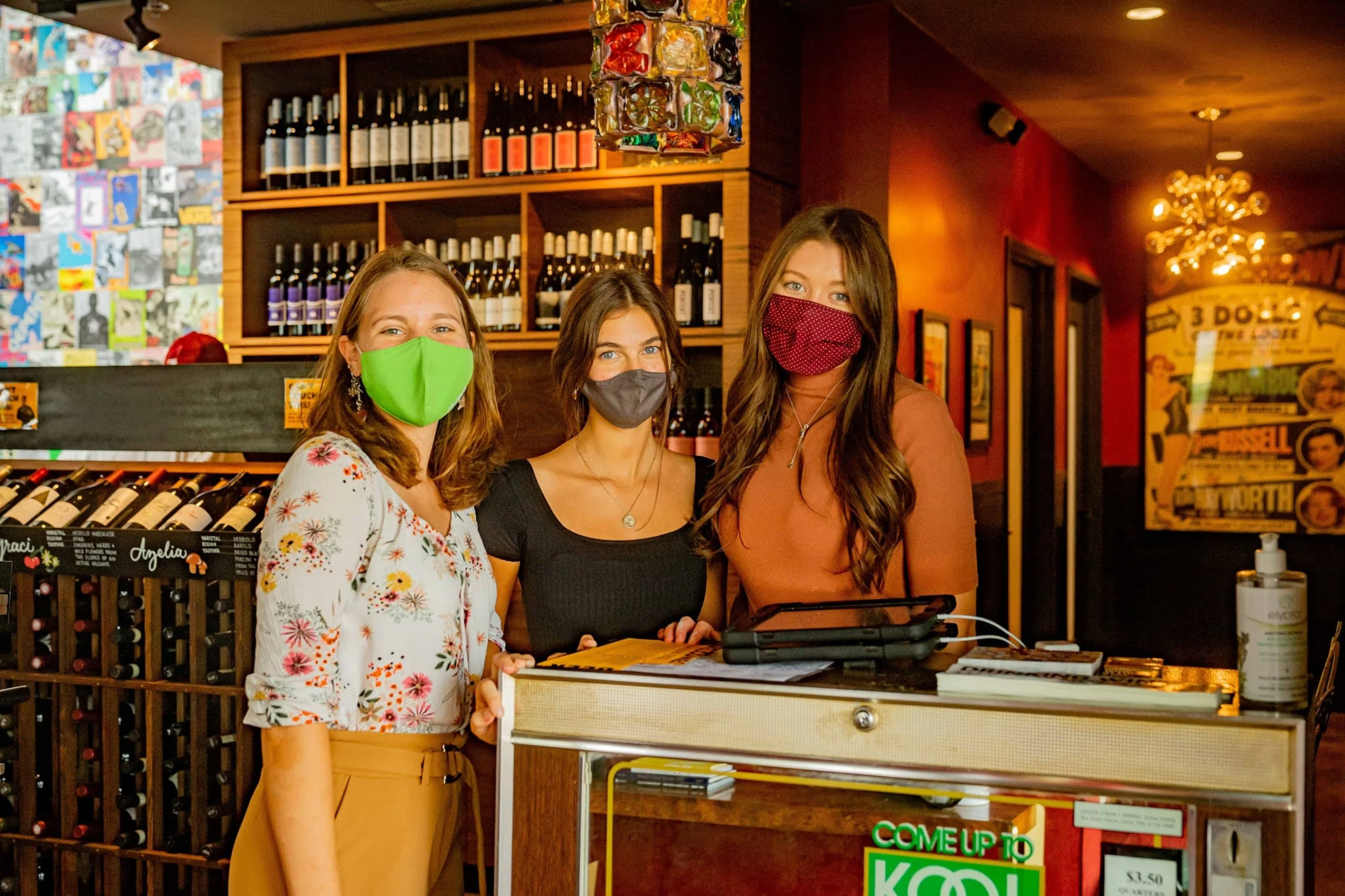 Three women wearing masks and posing near wine racks.