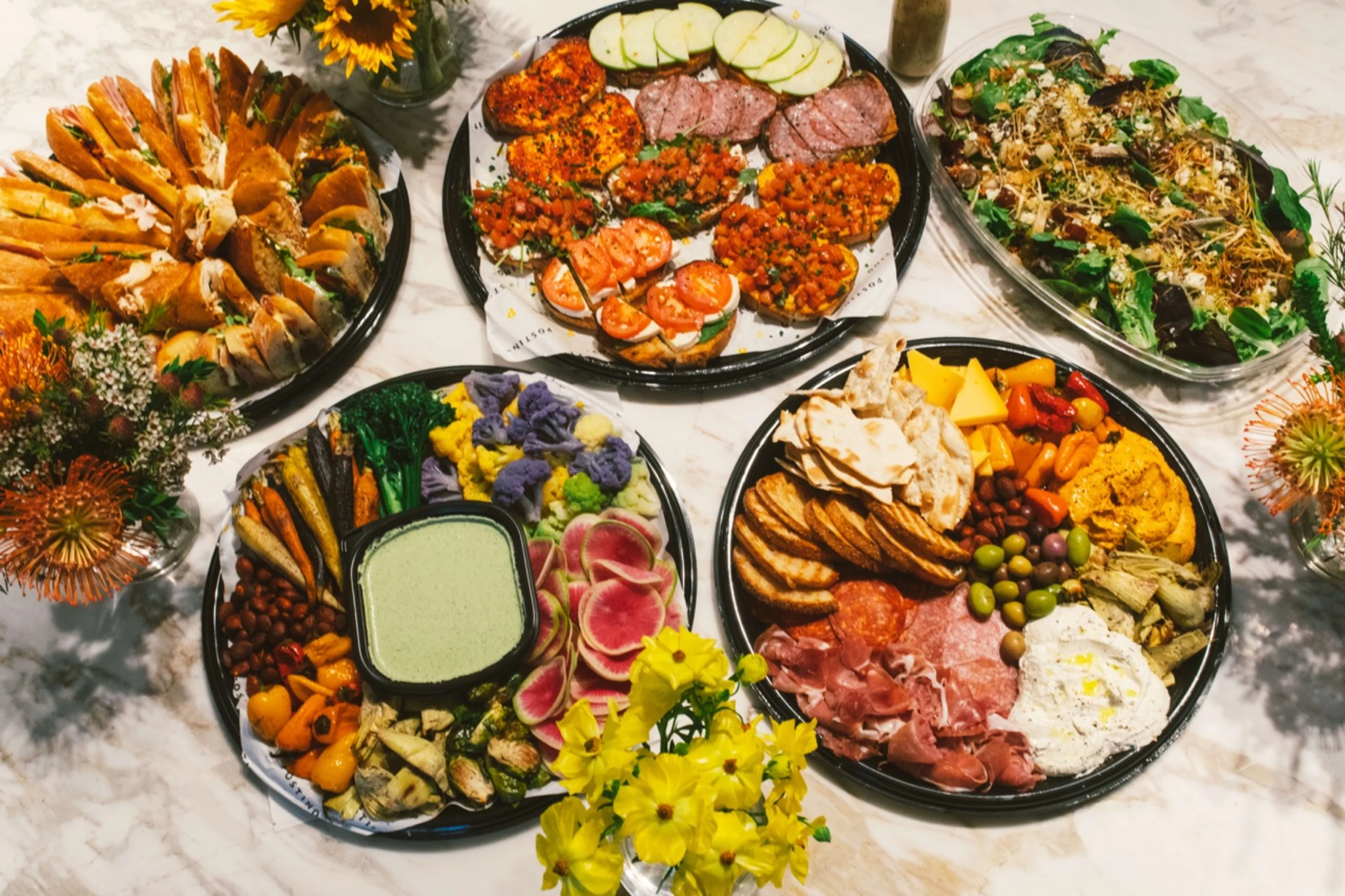 A table full of colorful food with three servers and a patron drinking beer.