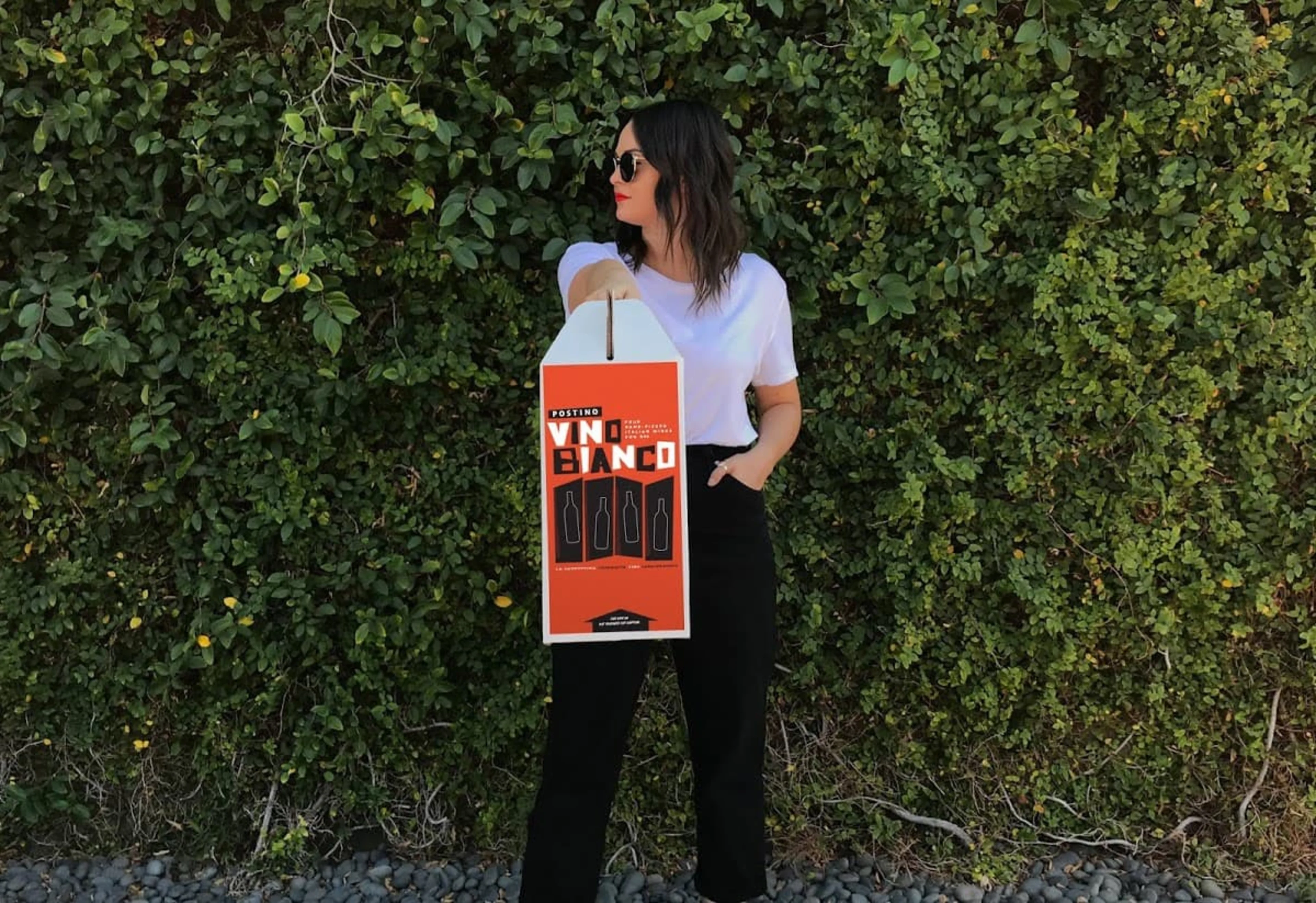 A woman holds a Vino Bianco orange box in front of a green shrub.