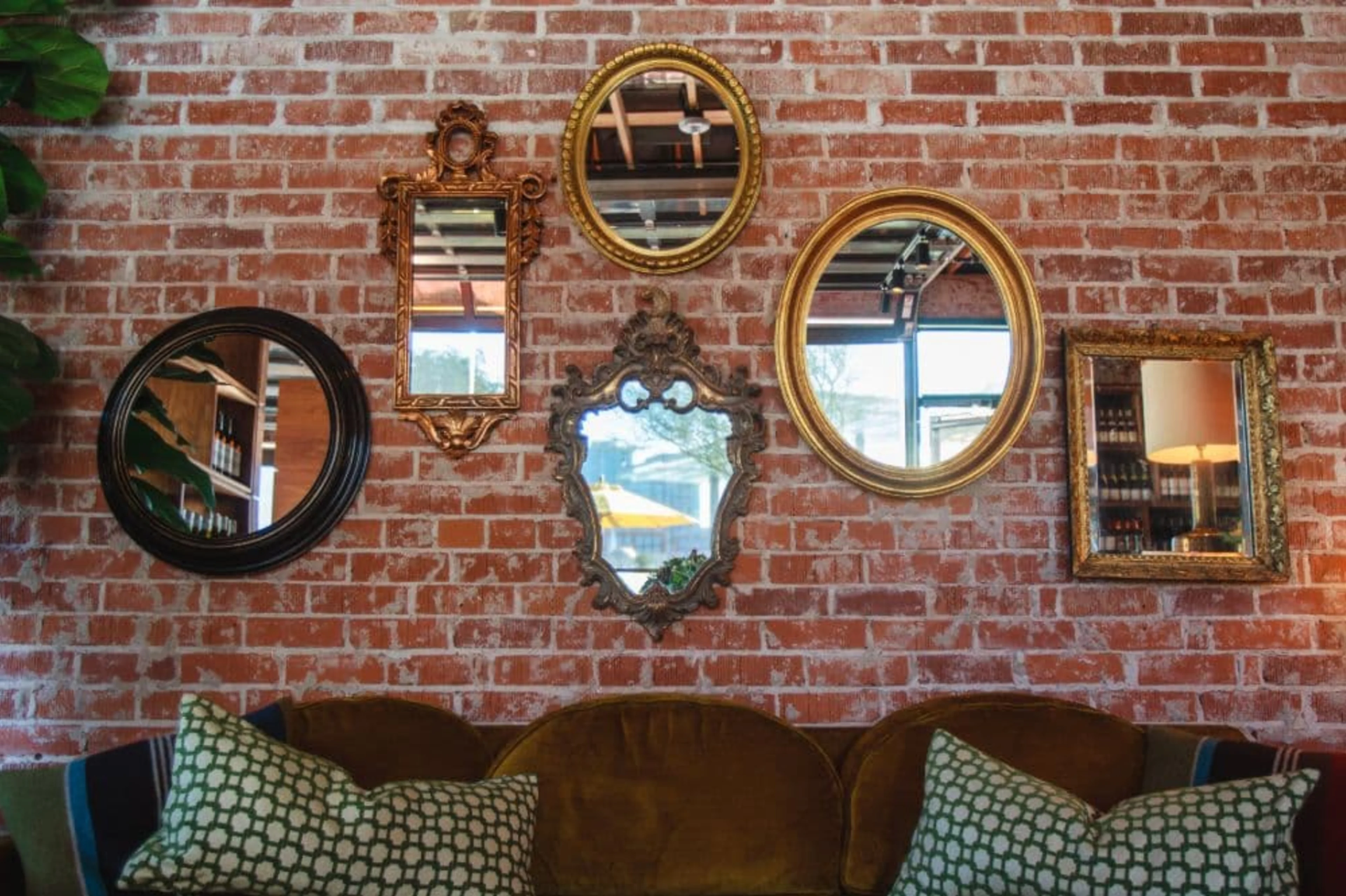 Brick wall with six mirrors mounted on it with a large brown sofa below. 