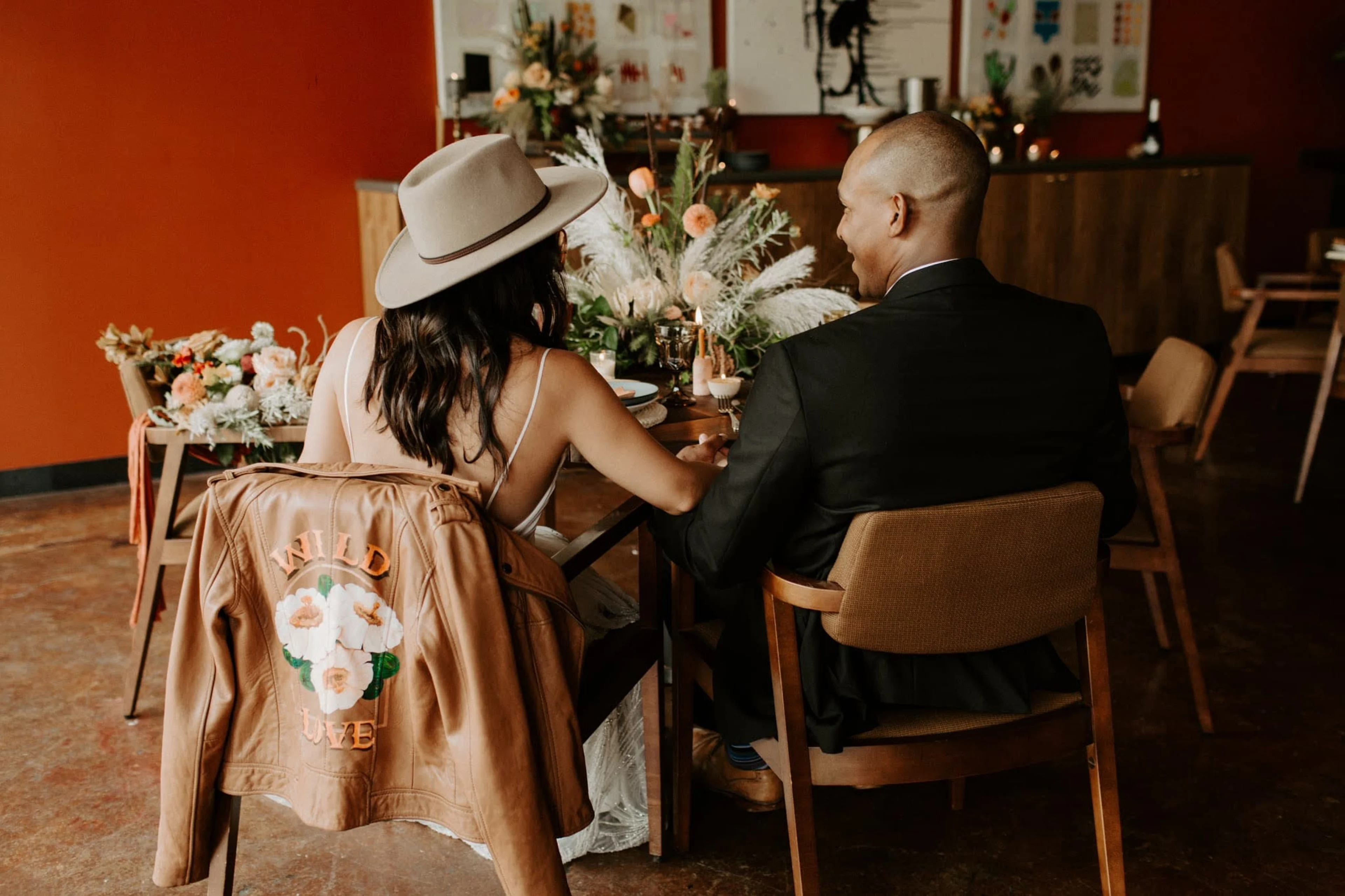 A bride and groom sit at a table with a jacket draped over a chair.