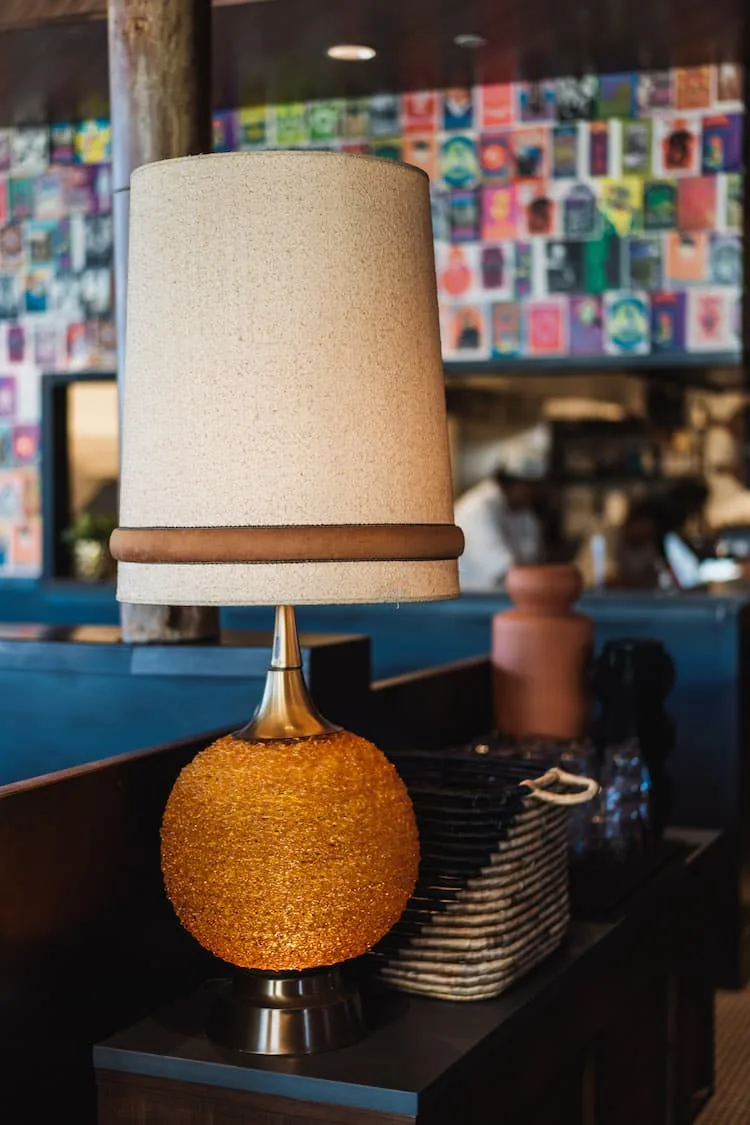 A brown and tan lamp on a table with a colorful wall behind it.