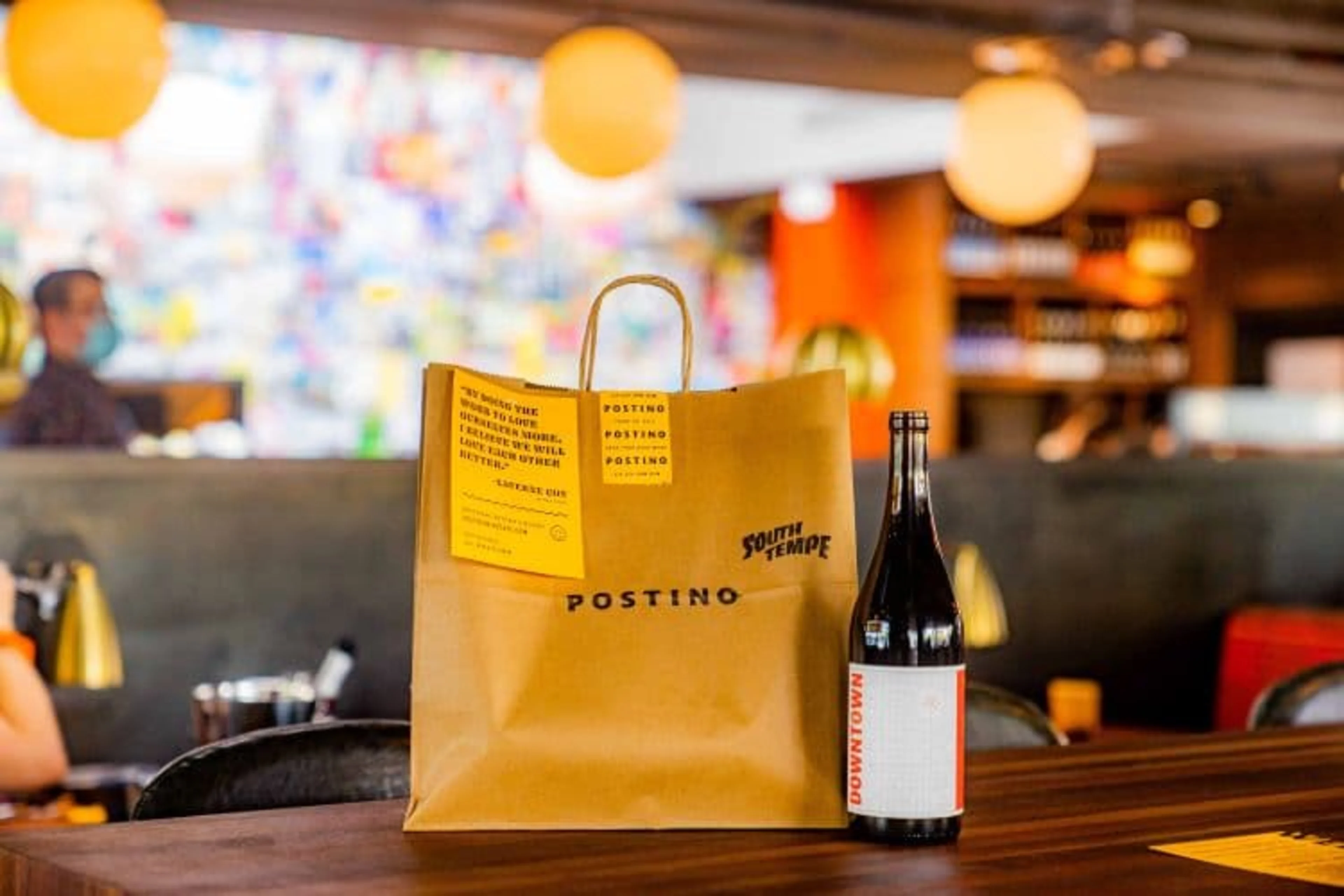 Postino to-go paper bag with a bottle of red wine next to it.
