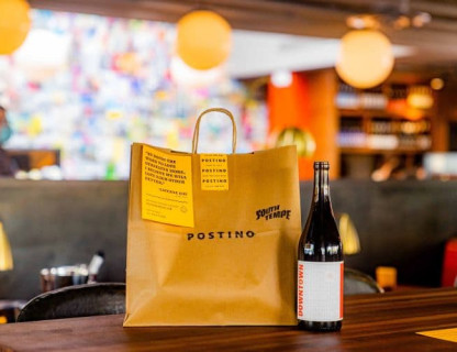 Postino to-go paper bag with a bottle of red wine next to it.
