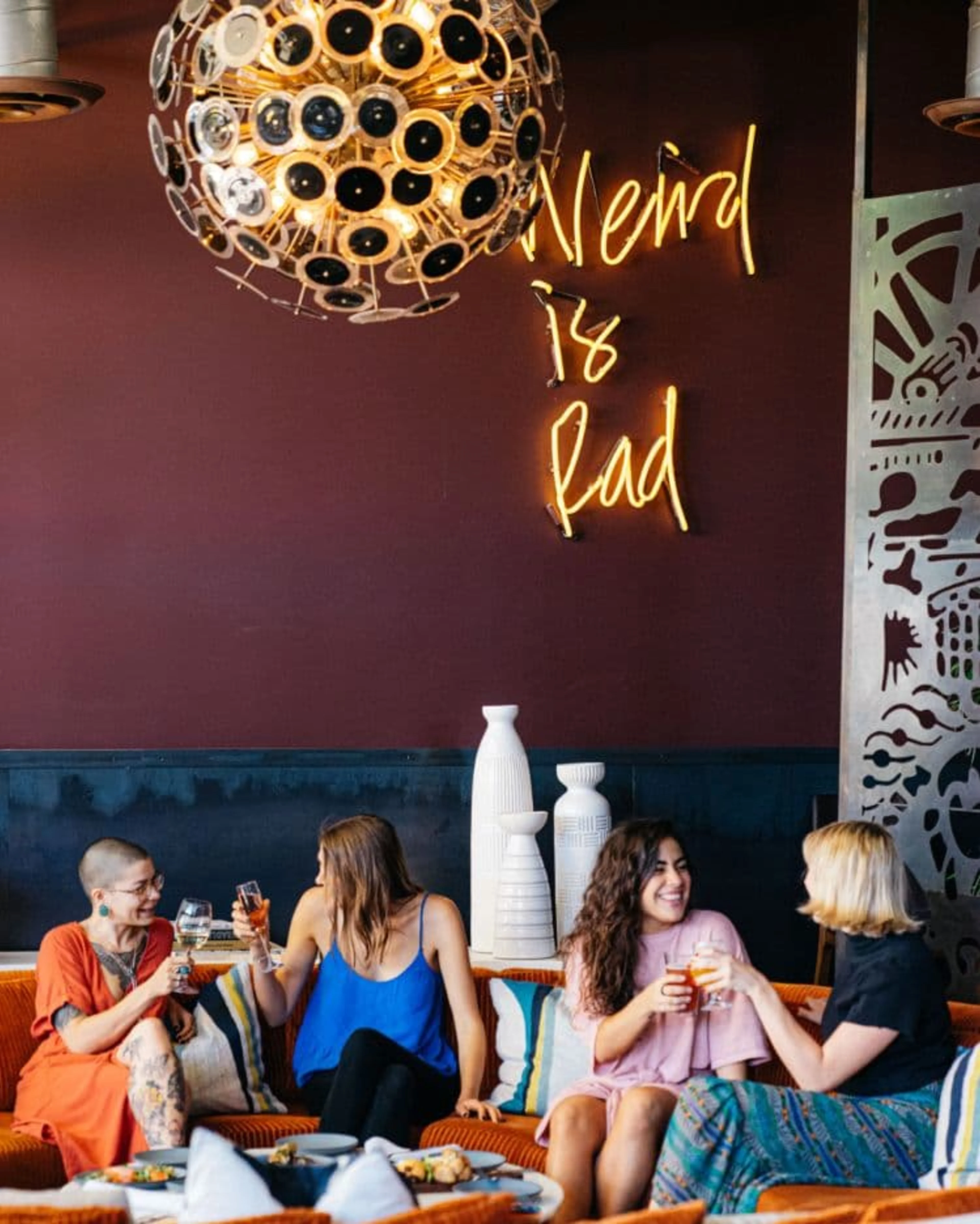 A burgundy wall with a sign "Weird is Rad" over four women clinking their wine glasses.