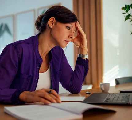 Person in purple shirt looking stressed while working on laptop at home desk with houseplant nearby.
