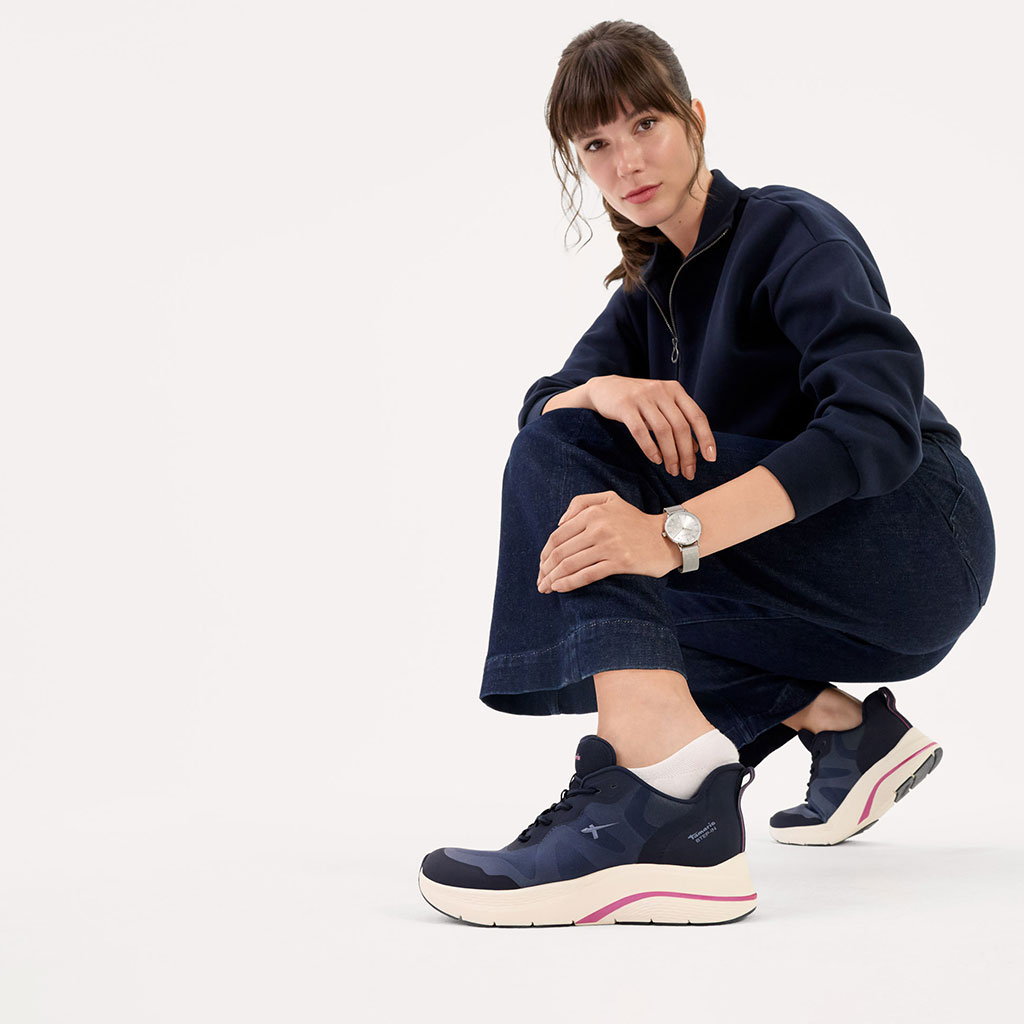 Une personne vêtue de vêtements de loisirs sombres et de chaussures de sport modernes pose accroupie sur un fond blanc.