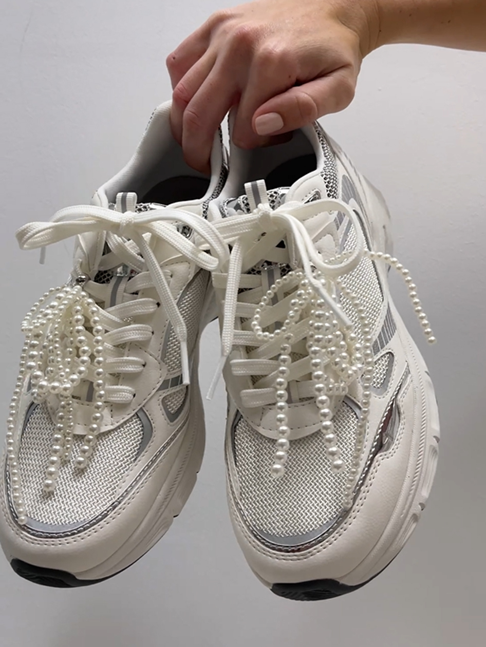 Hvide sneakers med perlebesatte sn&oslash;reb&aring;nd mod gr&aring; baggrund.