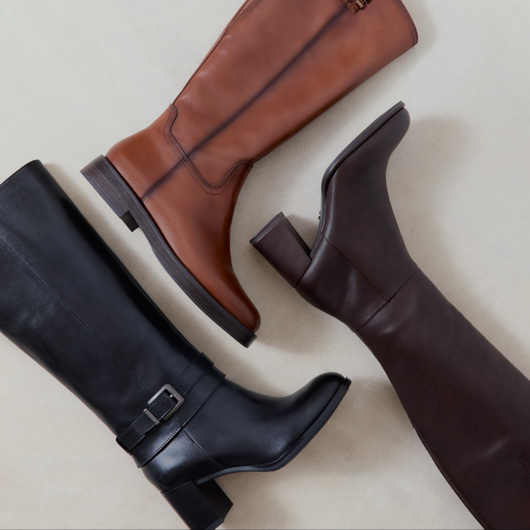 Drei elegante Lederstiefel in Schwarz, Cognac und Dunkelbraun auf hellem Hintergrund, flach fotografiert.