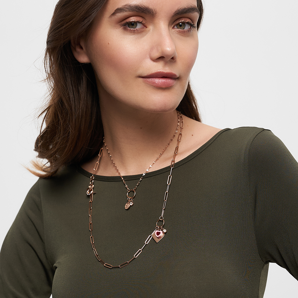 Gros plan sur un collier avec un pendentif en forme de cœur sur un haut vert olive, maquillage naturel et cheveux bruns au premier plan.