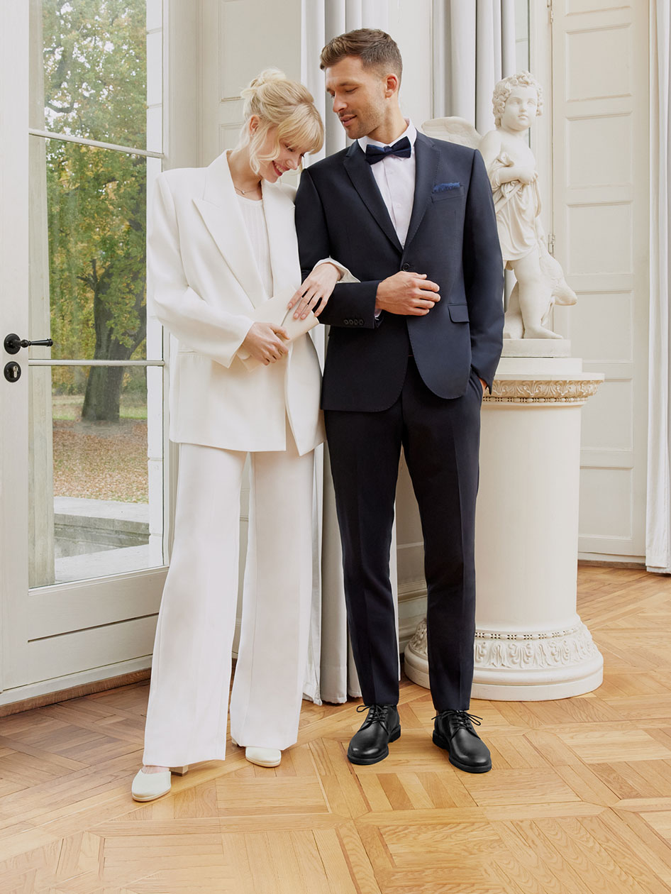 Elegantes Brautpaar in modernen Anz&uuml;gen - sie in Wei&szlig;, er in Schwarz - posiert in einem hellen Raum mit klassischer S&auml;ule und Parkblick.