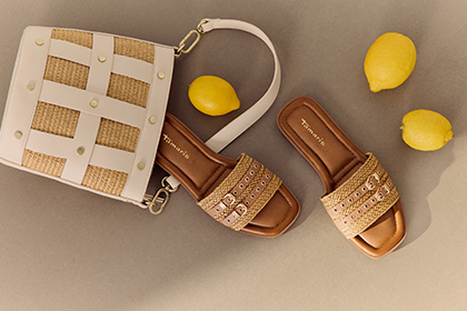 Braune Tamaris Sandalen und wei&szlig;-beige Flechthandtasche auf grauem Hintergrund, dekoriert mit gelben Zitronen.