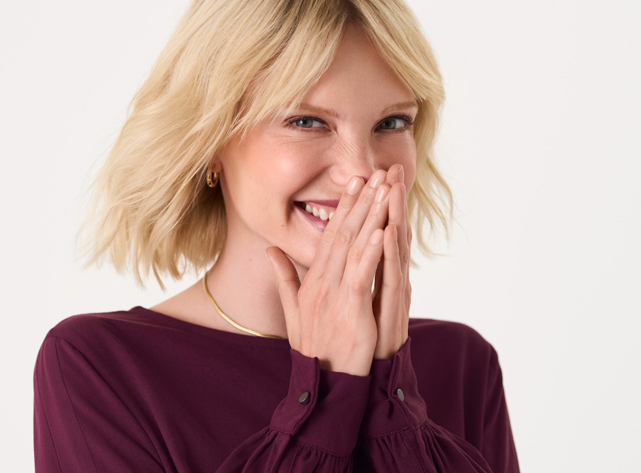 Nahaufnahme einer l&auml;chelnden Person mit blonden Haaren in burgunderroter Bluse, die verlegen ihre Hand vor den Mund h&auml;lt