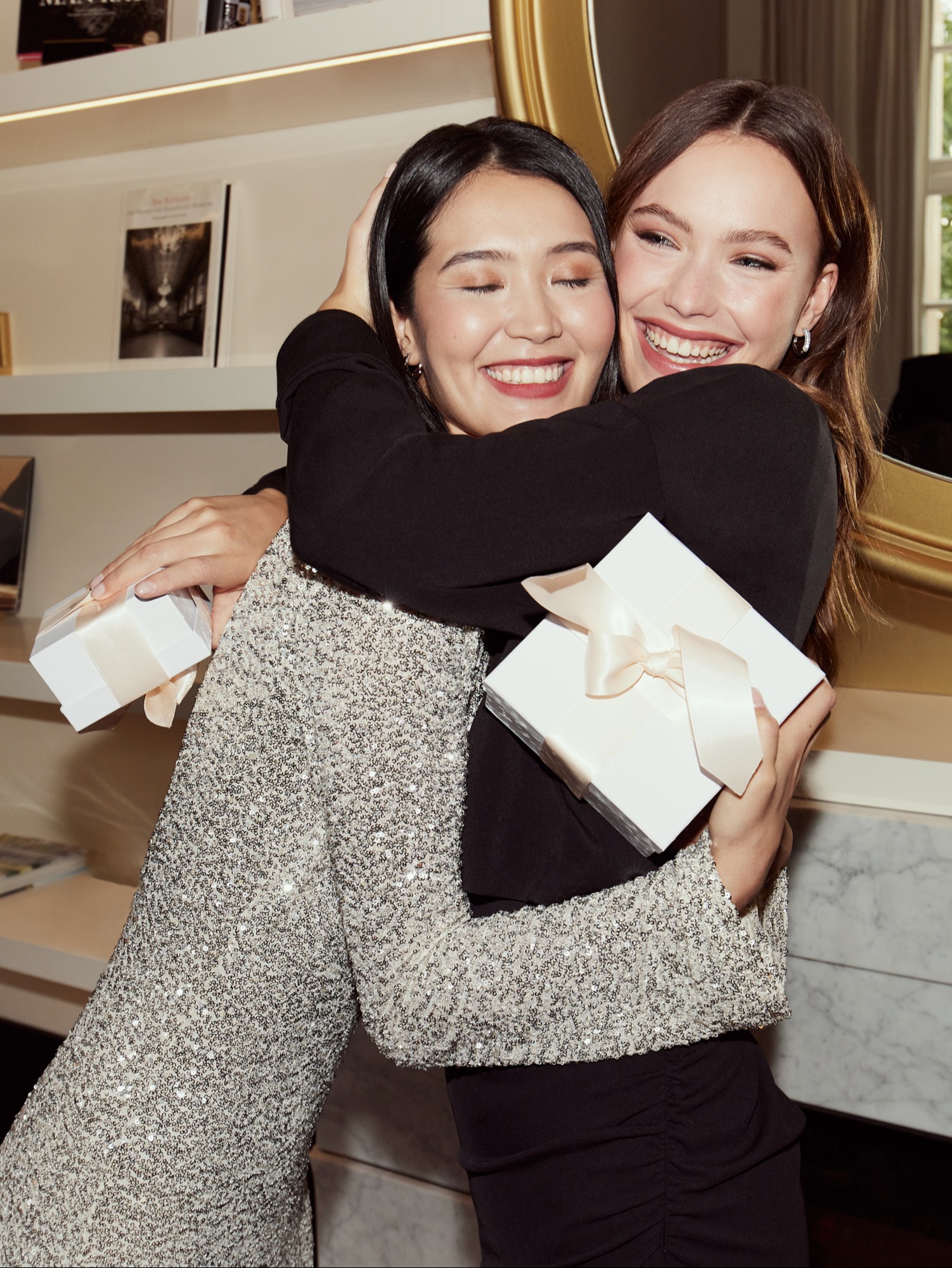 Zwei Freundinnen umarmen sich freudig, eine trägt ein glitzerndes Kleid, die andere ein schwarzes Outfit, beide halten weiße Geschenkboxen.