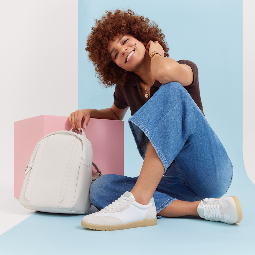 Femme en jean et t-shirt marron pose en souriant &agrave; c&ocirc;t&eacute; d&rsquo;un sac &agrave; dos blanc devant un fond pastel, portant des sneakers blanches.