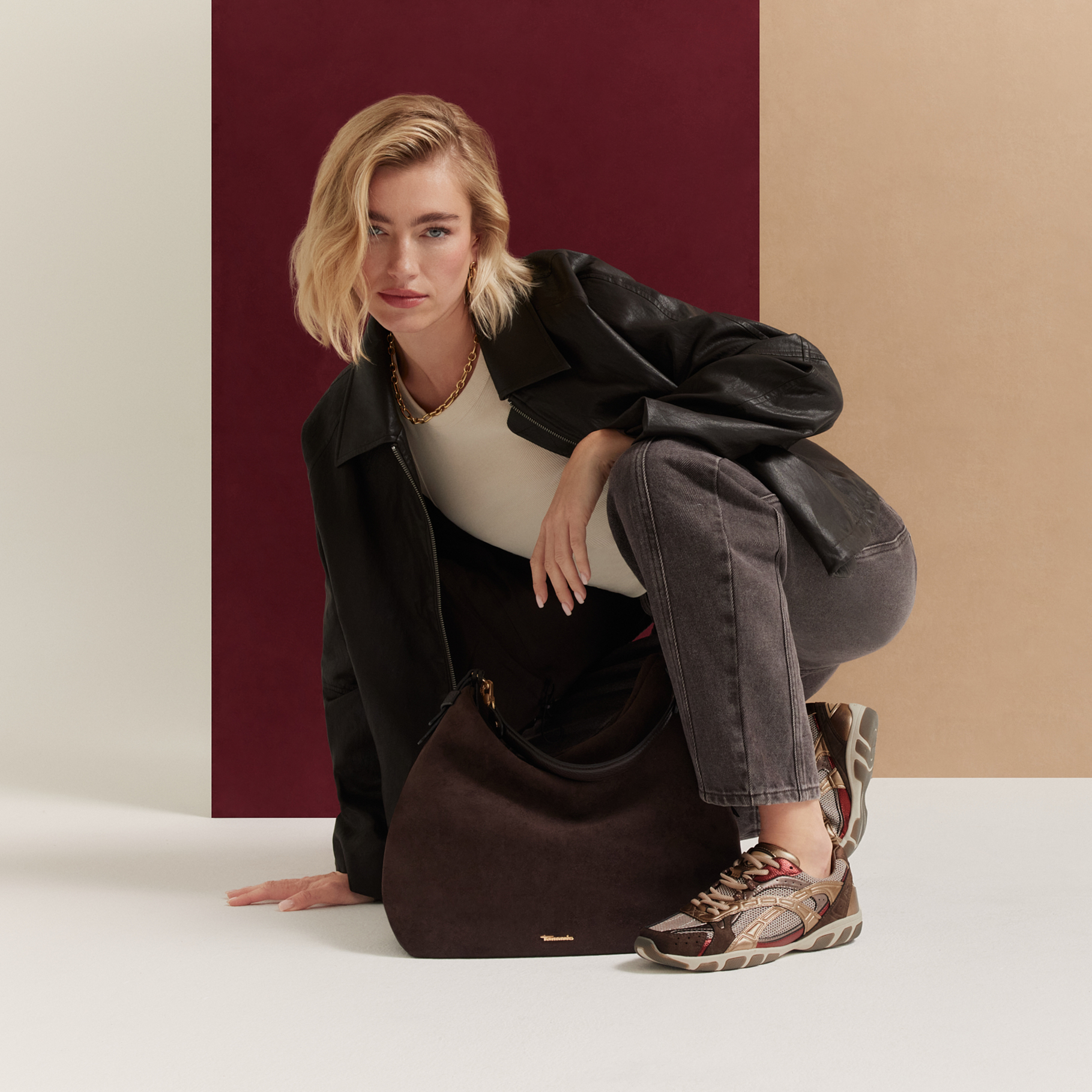 Woman in a black leather jacket crouching with a dark brown bag and colorful sneakers against a burgundy‑beige background.