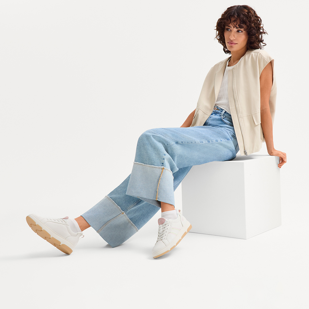Model in a beige vest, white T‑shirt and jeans sits on a white cube, wearing white sneakers with a beige sole.