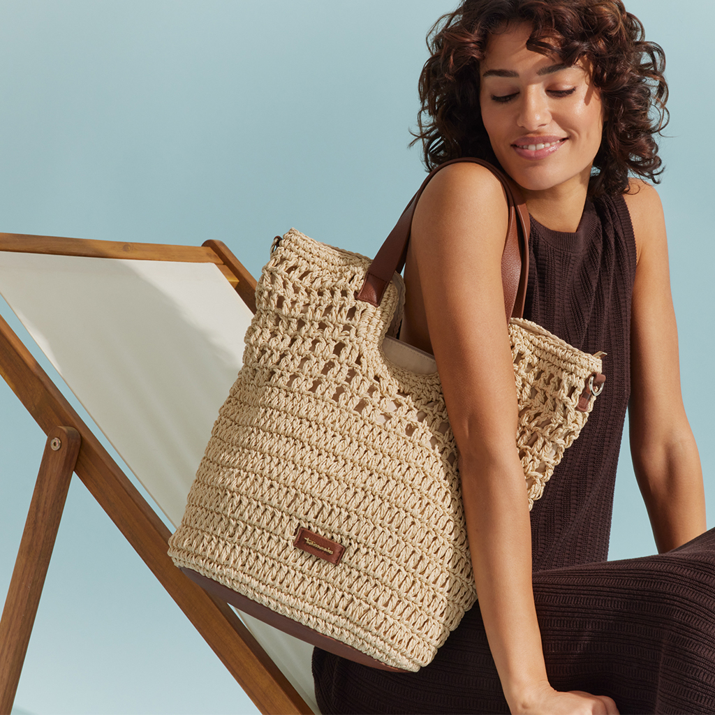 Vrouw in een donkerbruine jurk houdt een beige rieten tas met bruine leren hengsels en zit op een ligstoel.