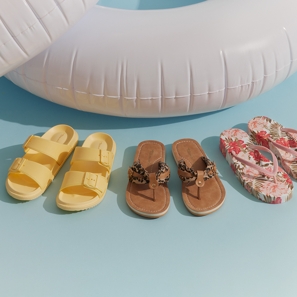 Drei Paar Sommerschuhe: gelbe Sandalen, braune Zehentrenner mit Leopardenmuster und rosa Flip-Flops mit Blumenmuster, neben einem wei&szlig;en Schwimmring.