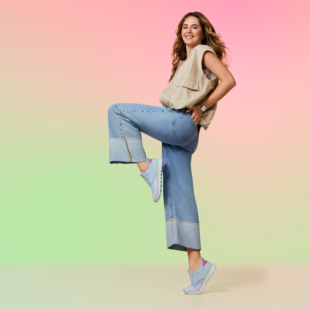 Kobieta w beżowej bluzce i dżinsach pozuje dynamicznie na pastelowym tle, mając na sobie jasnoniebieskie sneakersy z r&oacute;żowymi detalami.