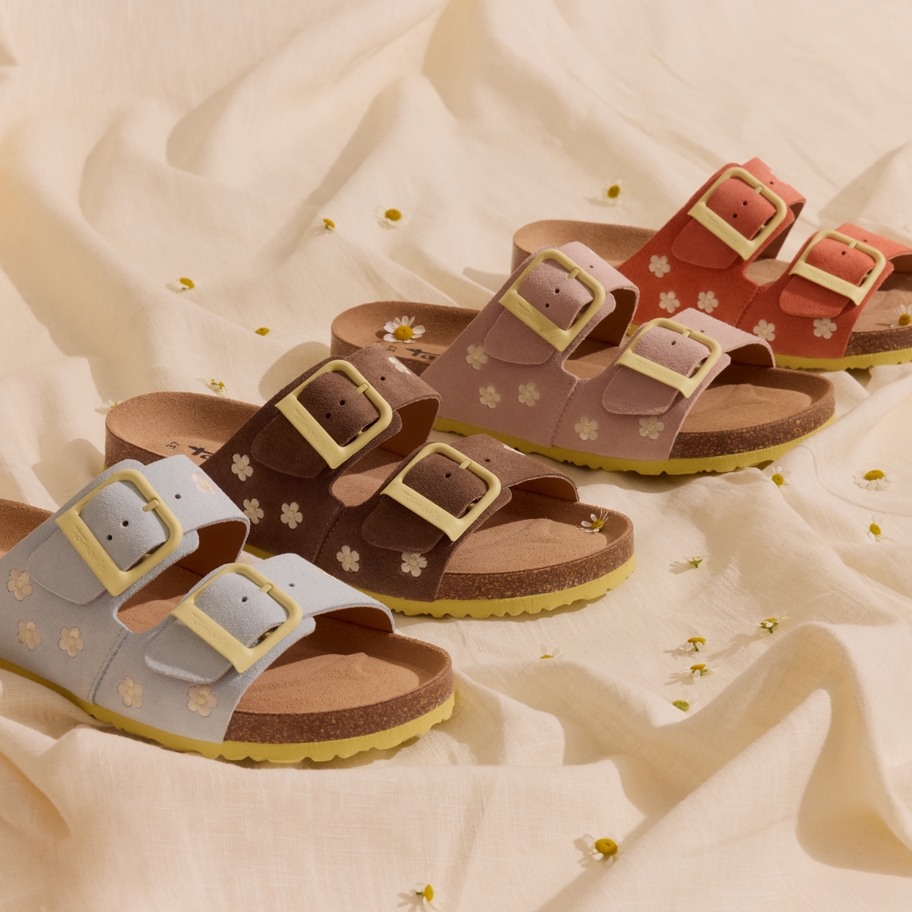 Mehrere Fu&szlig;bett-Sandalen in Pastellfarben mit Bl&uuml;tenstickerei, inszeniert auf beigem Leinenstoff mit kleinen G&auml;nsebl&uuml;mchen als Dekoration